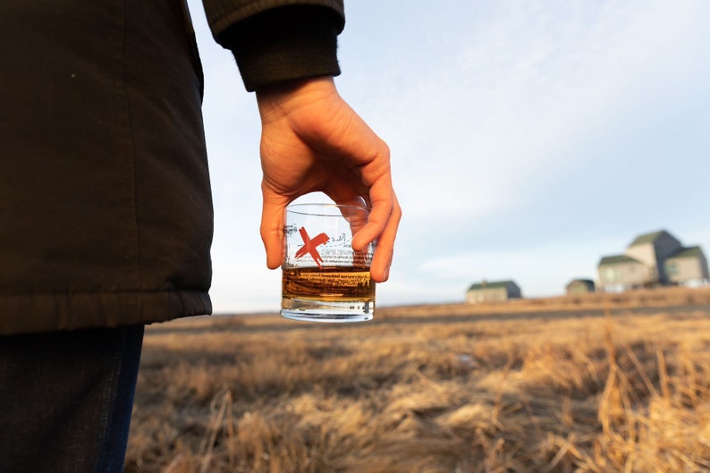 May include: A person is holding a glass of amber liquid with a red X on the side. The glass is in focus, while the person and the background are blurred. The background is a field of dry grass with a few buildings in the distance.