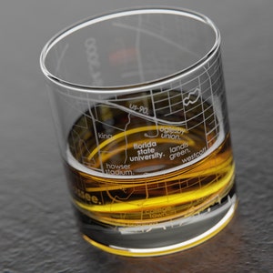 May include: A clear glass tumbler with a map of Tallahassee, Florida etched on the side. The glass is filled with a golden liquid, likely whiskey or bourbon. The map includes landmarks such as Florida State University, Howser Stadium, and Landis Green.