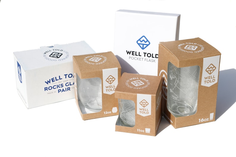 May include: Three brown cardboard boxes with clear glass cups inside. The boxes are labeled "Well Told" and have different sizes: 11oz, 12oz, and 16oz. A white box with the text "Well Told Pocket Flask" is also visible.