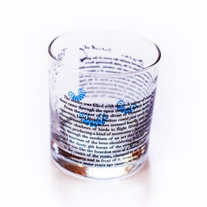 May include: A clear glass tumbler with a printed design featuring a quote about a studio filled with the rich odour of roses. The quote is printed in black text with blue accents.