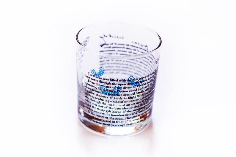 May include: A clear glass tumbler with a printed design featuring a quote about a studio filled with the rich odour of roses. The quote is printed in black text with blue accents.