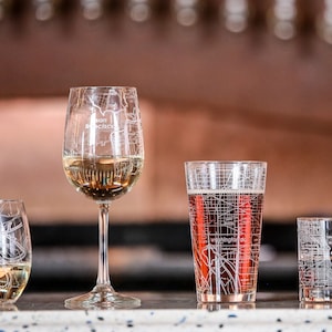 May include: Four glasses with city maps etched on them. The glasses are filled with different beverages. The glasses are on a counter top. The glasses are for wine, beer, and cocktails.