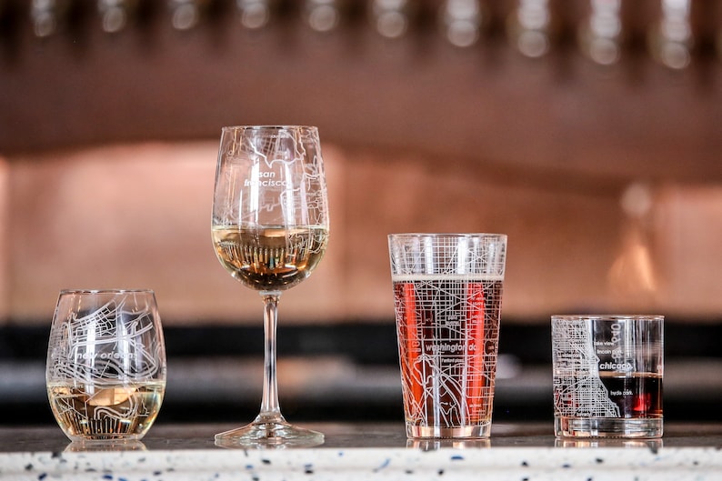 May include: Four glasses with city maps etched on them. The glasses are filled with different beverages. The glasses are on a counter top. The glasses are for wine, beer, and cocktails.