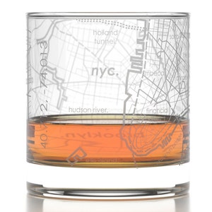 May include: A clear glass with a map of New York City etched on the side. The glass is filled with a brown liquid, likely whiskey or bourbon. The map includes the text "nyc." and "hudson river."
