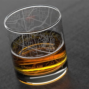 May include: A clear glass with an etched map of Chapel Hill, North Carolina, filled with a golden amber liquid. The glass has a rounded bottom and a wide mouth.