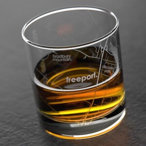 May include: A clear, cylindrical glass with a map design and the word "freeport" etched on the side. The glass is filled with a golden-brown liquid, possibly whiskey or a similar spirit. The glass is on a dark surface.