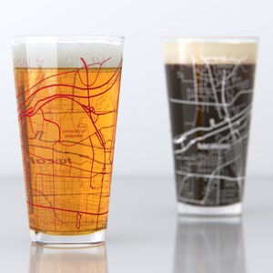 May include: Two pint glasses, one filled with beer and the other with a dark stout. Each glass features a map design. The beer glass has a red map, and the stout glass has a white map. The beer glass has the text "University of Alabama".