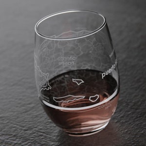May include: A clear glass wine glass with an etched map of Puerto Rico. The glass is filled with a pink beverage. The text "atlantic ocean." is etched on the glass.