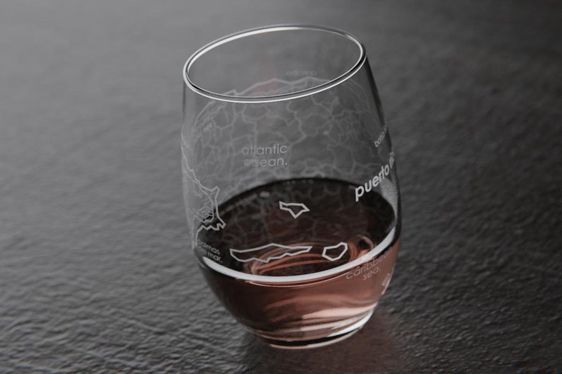 May include: A clear glass wine glass with an etched map of Puerto Rico. The glass is filled with a pink beverage. The text "atlantic ocean." is etched on the glass.