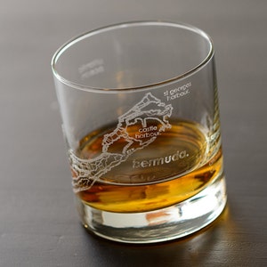 May include: A clear glass with a brown liquid inside. The glass has an etched design of the island of Bermuda with the words "Bermuda", "St. George's Harbour", and "Castle Harbour" on it.