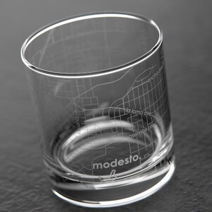 May include: A clear glass tumbler with an etched design of the city of Modesto, California. The design includes street names and the word "modesto" at the bottom.