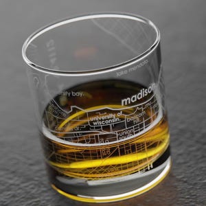 May include: A clear glass with a map of Madison, Wisconsin etched on the side. The glass is filled with a golden liquid.