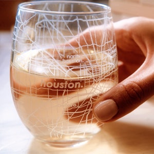 May include: A clear glass wine glass with a white outline of the Houston, Texas street map. The glass is filled with a light-colored beverage.