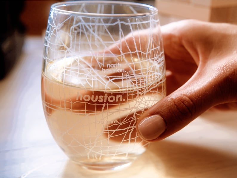 May include: A clear glass wine glass with a white outline of the Houston, Texas street map. The glass is filled with a light-colored beverage.