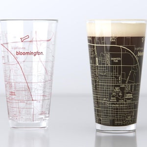 May include: Two clear glass pint glasses with a map of Bloomington, Indiana printed on them. The glass on the left is empty, while the glass on the right is filled with a dark brown liquid.