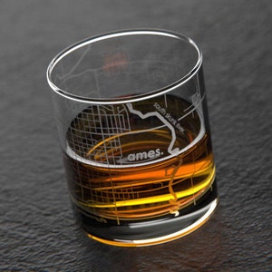 May include: A clear glass with a design of the city of Ames, Iowa, etched into the glass. The glass is filled with amber-colored liquid. The text "south skunk river." is etched into the glass.