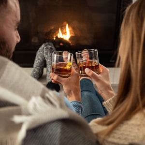 May include: A couple relax by a fireplace, each holding a glass of amber liquid. They are wrapped in blankets and seem to be enjoying a cosy evening.