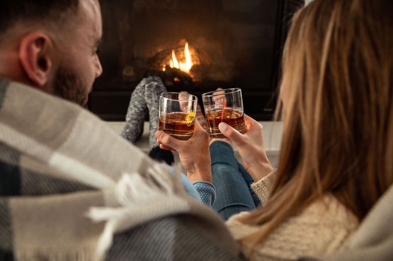 May include: A couple relax by a fireplace, each holding a glass of amber liquid. They are wrapped in blankets and seem to be enjoying a cosy evening.