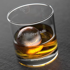 May include: A clear, lowball whiskey glass with a spherical ice ball inside, filled with amber liquid. The glass has the phrase "kill devil hills." etched on the side. The glass is on a dark gray surface.