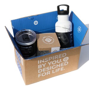 May include: A cardboard box with the words "Inspired by you. Designed for life." printed on the side. The box contains a white water bottle with a black lid and a black and silver tumbler. The box also contains a small brown box with the words "Well Told Life" printed on it.