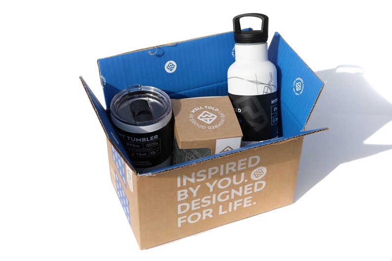 May include: A cardboard box with the words "Inspired by you. Designed for life." printed on the side. The box contains a white water bottle with a black lid and a black and silver tumbler. The box also contains a small brown box with the words "Well Told Life" printed on it.