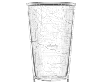 May include: A clear glass pint glass etched with a map of Atlanta, Georgia. The city's name is printed in a simple font. The map includes street names and neighborhood labels. The glass is cylindrical with a slightly tapered shape.