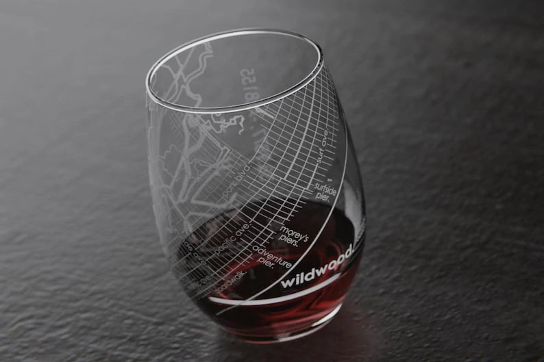May include: A clear glass wine glass with a design etched into the side. The design features a map of Wildwood, New Jersey, with various landmarks and streets labeled. The glass is filled with red wine.