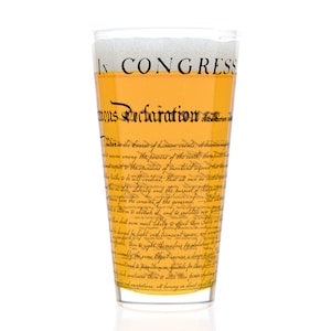 May include: A tall glass filled with a light yellow liquid, with a white head of foam. The glass is printed with the text "In Congress, July 4, 1776.  Unanimous Declaration of the thirteen united States of America."
