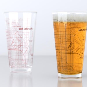 May include: Two clear glass pint glasses with a map of Salt Lake City printed on them. One glass is empty and the other is filled with a light amber colored beverage.