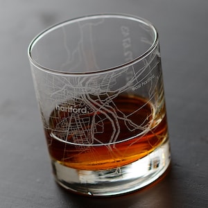 May include: A clear glass tumbler etched with a map of Hartford, Connecticut, filled with amber liquid. The city's name is visible on the glass. The glass sits on a dark surface.