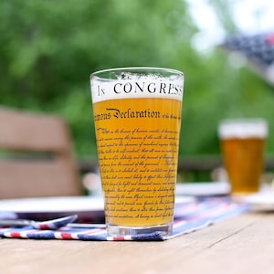 May include: A glass of beer with the text "In Congress, July 4, 1776. Unanimous Declaration" printed on the side. The glass is sitting on a wooden table with a red, white, and blue tablecloth.