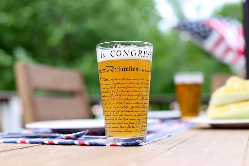 May include: A glass of beer with the text "In Congress, July 4, 1776. Unanimous Declaration" printed on the side. The glass is sitting on a wooden table with a red, white, and blue tablecloth.