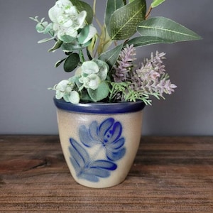 Puede incluir: Una pequeña maceta de cerámica de color crema con un diseño floral azul. La maceta está llena de una variedad de plantas artificiales verdes y blancas.