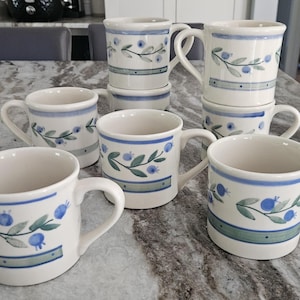 May include: A set of white ceramic mugs featuring a blue and green floral pattern. Each mug has a handle and a blue band around the top and bottom. The design includes blue berries and green leaves.
