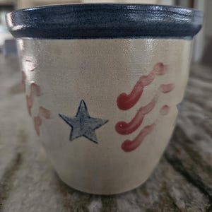 May include: A ceramic pot with a blue rim and a cream-colored body. The pot is decorated with a blue star and red wavy lines, reminiscent of a patriotic theme. The pot is likely used for holding items.