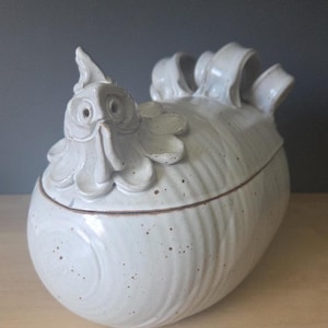 May include: A white ceramic chicken-shaped container with a lid. The container has a detailed head with eyes, a beak, and a comb. The body is rounded with textured details. The lid has decorative elements. The ceramic piece is speckled with brown spots.