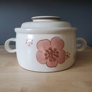 May include: A white ceramic casserole dish with a lid, featuring a pink floral design with brown accents. The dish has two handles on the sides.