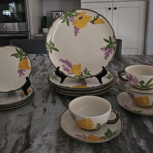 Puede incluir: Un juego de vajilla vintage con estampado floral, que incluye platos y tazas de t&eacute;. Los platos presentan un dise&ntilde;o floral amarillo y morado con hojas verdes. Las tazas de t&eacute; tienen un fondo blanco con un dise&ntilde;o floral amarillo y morado.