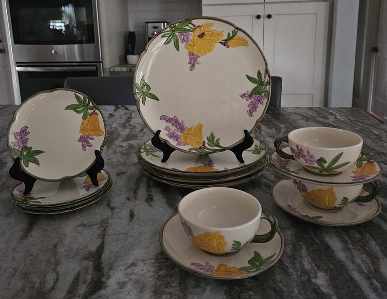 Puede incluir: Un juego de vajilla vintage con estampado floral, que incluye platos y tazas de t&eacute;. Los platos presentan un dise&ntilde;o floral amarillo y morado con hojas verdes. Las tazas de t&eacute; tienen un fondo blanco con un dise&ntilde;o floral amarillo y morado.