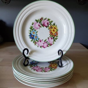 May include: Set of six white ceramic dinner plates with a green rim and a floral design in the center. The floral design is a wreath of pink, yellow, blue, and green flowers.