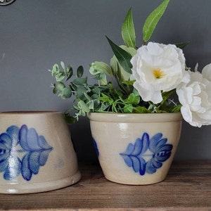 May include: Two ceramic planters with blue and white floral designs. The planter on the right is filled with white flowers and green leaves.