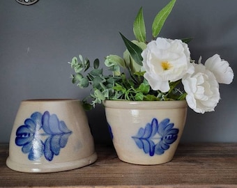 Puede incluir: Dos macetas de cerámica con diseños florales azules y blancos. La maceta de la derecha está llena de flores blancas y hojas verdes.