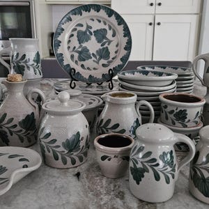 May include: A collection of handmade pottery in shades of gray and green with a leafy design. The pottery includes mugs, pitchers, bowls, and a serving dish.