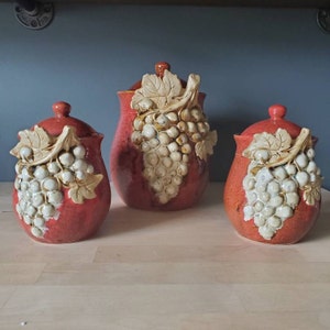 Könnte beinhalten: Set aus drei roten Keramikbehältern mit weißen Traubenmotiven. Die Behälter haben Deckel und sind mit einem Weinrebenmotiv verziert.