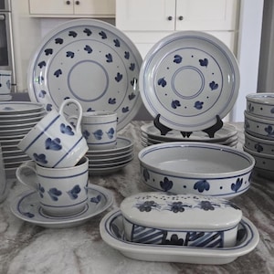 May include: A ceramic dinnerware set featuring a white base with blue hand-painted floral patterns. The collection includes a large pitcher, dinner plates, salad plates, cups with saucers, a butter dish with a lid, and several deep bowls.