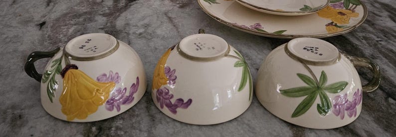 Puede incluir: Tres tazas de t&eacute; de cer&aacute;mica blanca con dise&ntilde;os florales. Las tazas presentan flores amarillas, moradas y verdes sobre un fondo blanco. Las tazas son todas ligeramente diferentes, pero tienen el mismo dise&ntilde;o general.