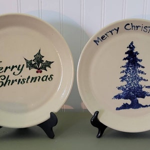 May include: Two white ceramic plates with black stands. The plate on the left has a green holly leaf design and the text "Merry Christmas". The plate on the right has a blue tree design and the text "Merry Christmas".