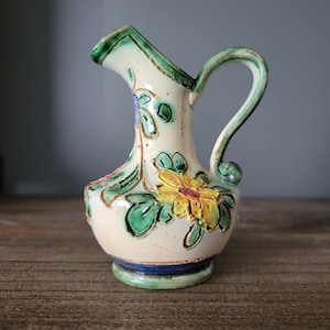 Könnte beinhalten: Ein kleiner, weißer Keramik Krug mit einem grünen Henkel und Blumendesigns in den Farben Gelb, Grün, Blau und Rot.