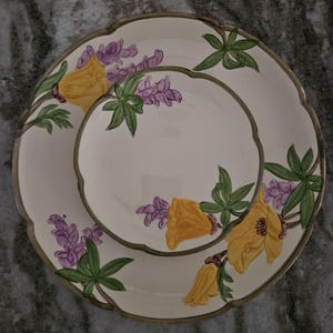 Puede incluir: Un juego de dos platos de cer&aacute;mica blanca con un dise&ntilde;o floral que presenta flores amarillas y flores de lupino p&uacute;rpura. Los platos tienen un borde festoneado y un borde verde.