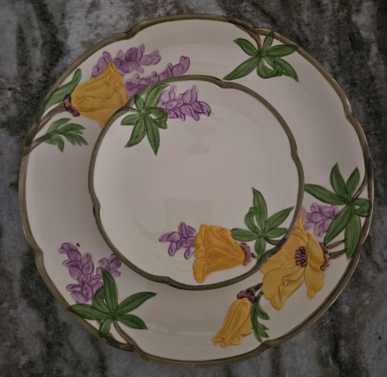 Puede incluir: Un juego de dos platos de cer&aacute;mica blanca con un dise&ntilde;o floral que presenta flores amarillas y flores de lupino p&uacute;rpura. Los platos tienen un borde festoneado y un borde verde.